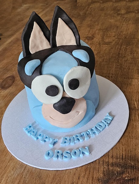 A round cake modelled as a blue cartoon dog’s face, lettered Happy Birthday Orson