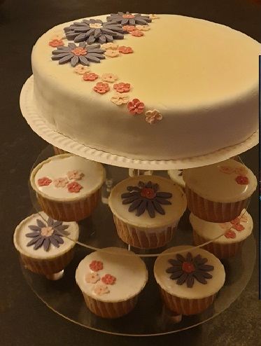 An ivory iced cake on a stand with matching daisy-topped cup cakes below