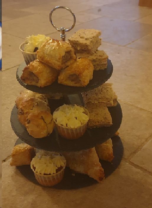 A three-tier slate stand of sausage rolls, sandwiches, cup cakes and pasties